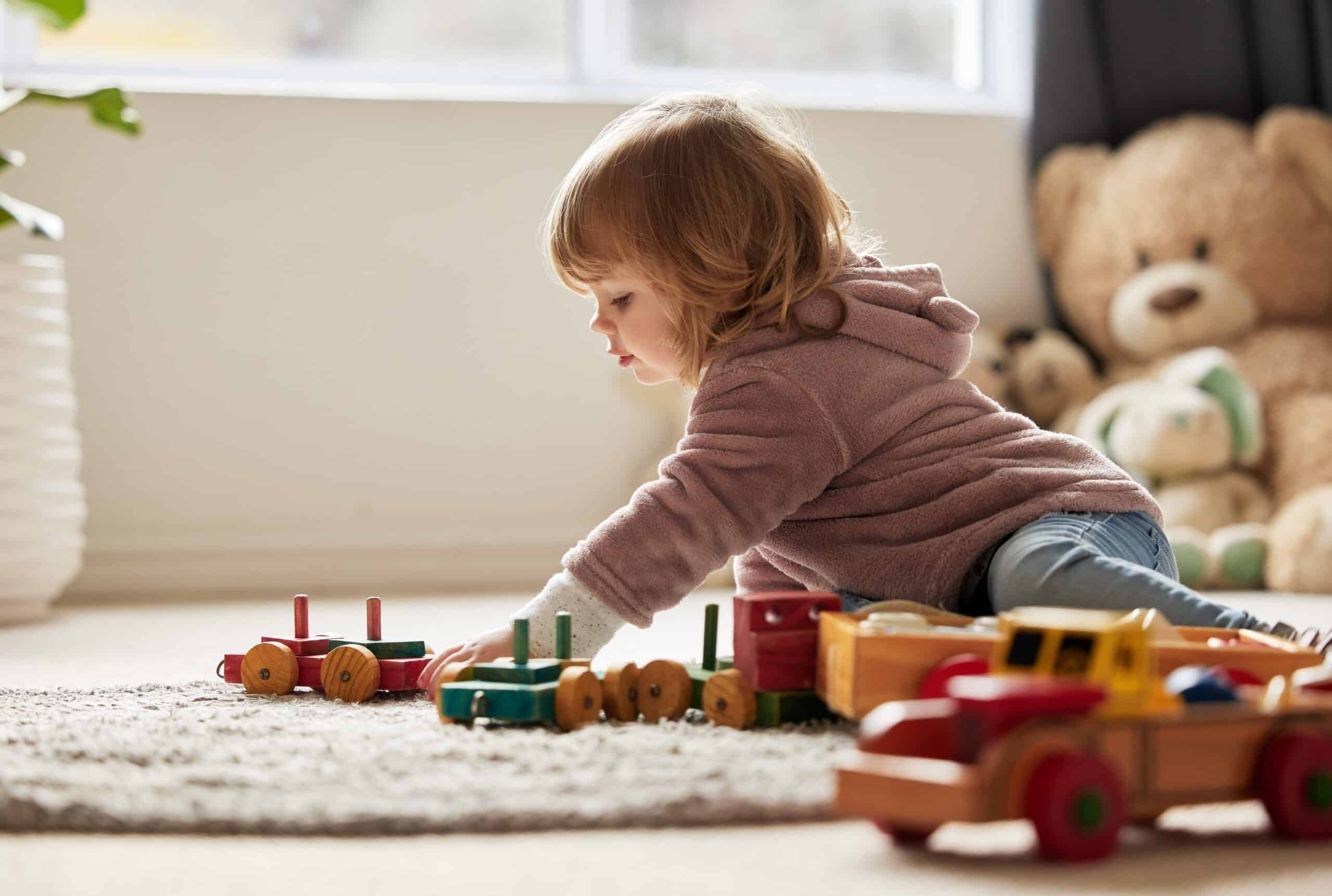 Toddler playing with wooden toy trains on a rug near a teddy bear