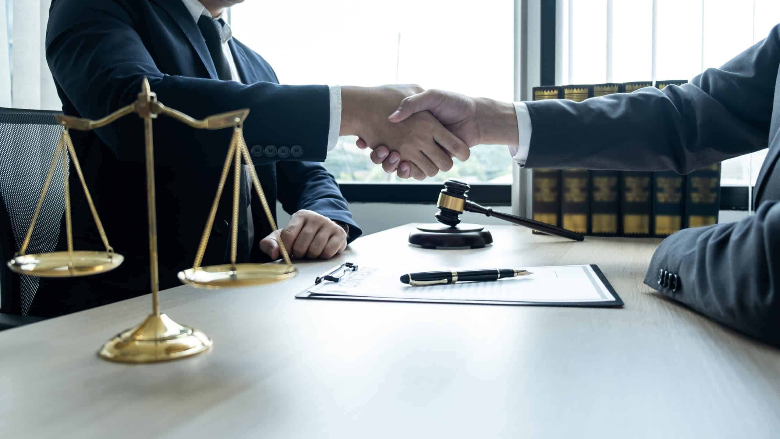 Two people shaking hands across a desk beside scales of justice and a gavel