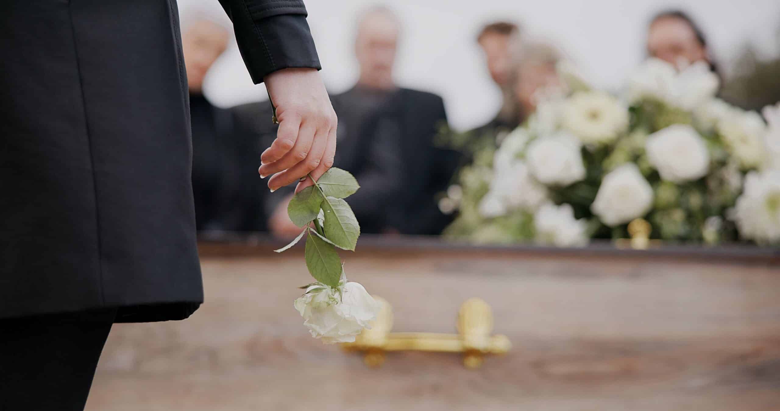 Hand placing a white rose on a casket as mourners gather