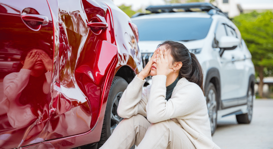 Distressed woman holding her face beside a red car with rear-end damage