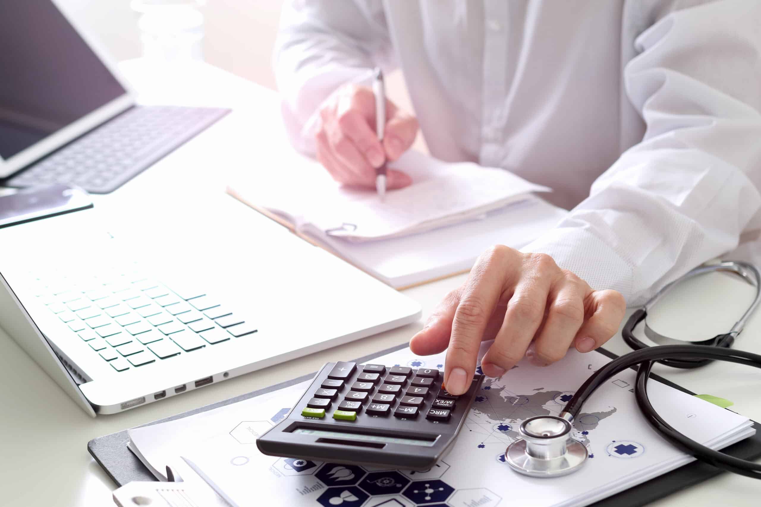 Hands using a calculator and laptop beside a stethoscope to total medical bills