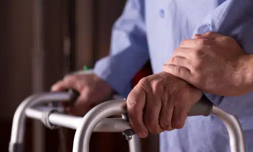 An elderly man's hands grasping the handles of a walking aid while being assisted by another person.
