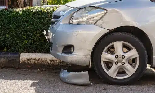 A low shot of a silver car with a part of its front bumper torn off after an accident.