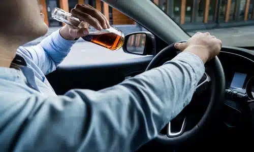 A driver with a bottle of alcohol in hand while operating a vehicle in the middle of the day.