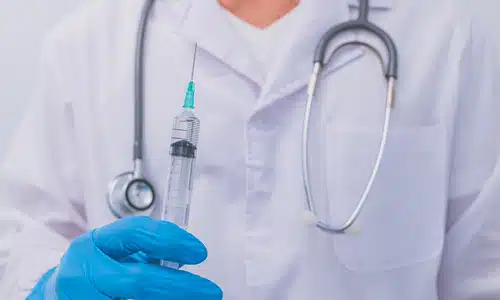 A closeup of a physician holding a up a syringe with a gloved hand at chest level.