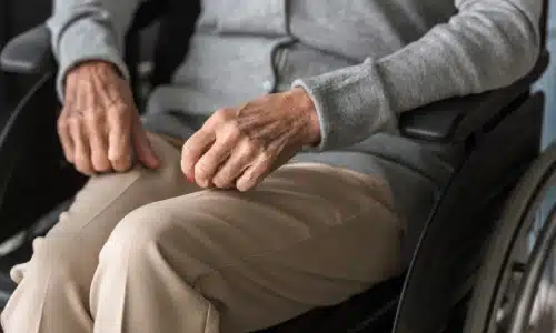 A closeup of an elderly woman seated in a wheelchair with her hands on the armrests.
