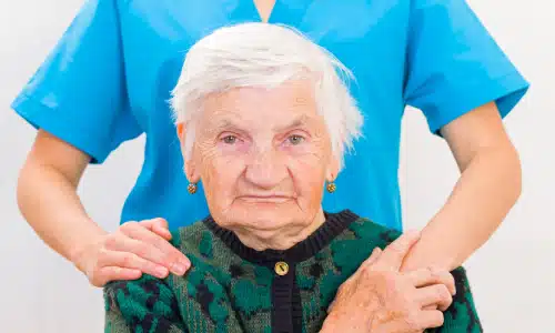 An elderly woman seated and staring at the viewer while a caregiver stands behind her with arms on her shoulders.