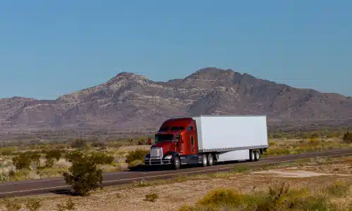 A red semi truck on a plains road pulling a large, white trailer behind it.