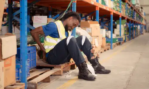 An injured warehouse worker sitting down on a pallet and holding his hip in pain.