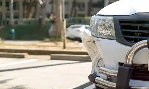 A closeup shot of a damaged passenger-side bumper of a white vehicle stopped at the side of a city road.