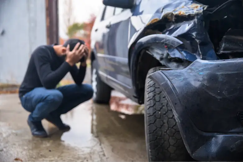 A distressed man crouching beside the crushed fender of his vehicle after a car accident.