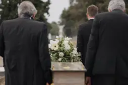 Mourners in black suits carrying a coffin with white flowers, representing a solemn scene of a wrongful death funeral procession.