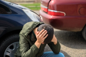 A car accident victim with his head resting on his hands while he sits next to the front wheel of his vehicle.