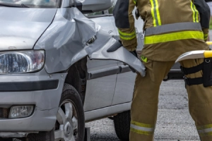 A pair of firefighters attempting to open the driver's side door of a grey vehicle at a car accident scene.
