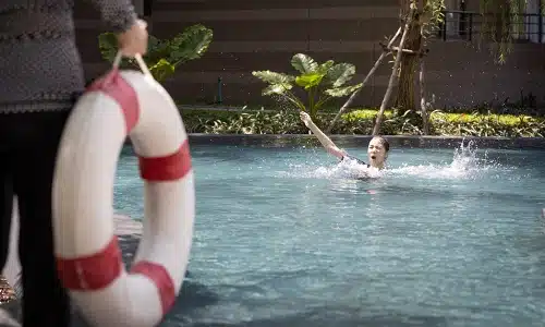 A man holding a lifesaver next to a pool about to throw it to a drowning woman.